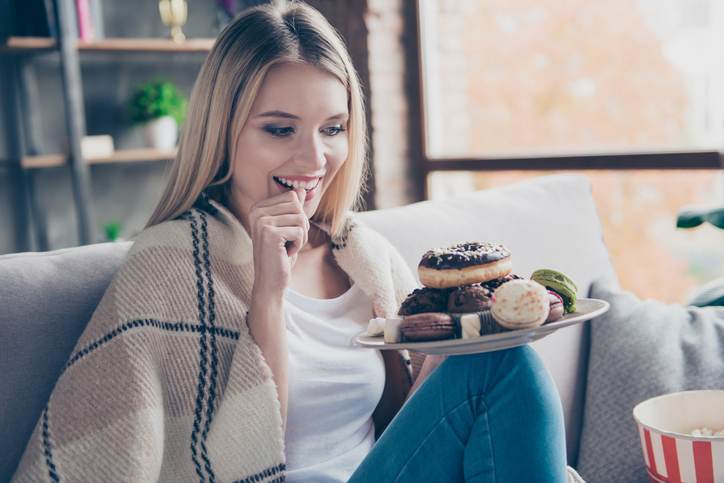 Portrait of beautiful emotional charming attractive sweet toothy woman sitting on sofa in living room, holding plate of donuts and macaroons, looking exciting satisfied